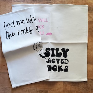 May include: Three off-white kitchen towels with black and pink text. One towel reads "find me where the rocks at". Another says "WILL ROCK U!" with a pink cloud. The third says "SILY BACTED OCK" with a speech bubble that says "I GOT A ROCK".