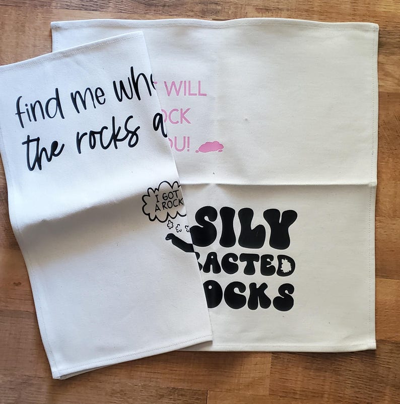 May include: Three off-white kitchen towels with black and pink text. One towel reads "find me where the rocks at". Another says "WILL ROCK U!" with a pink cloud. The third says "SILY BACTED OCK" with a speech bubble that says "I GOT A ROCK".