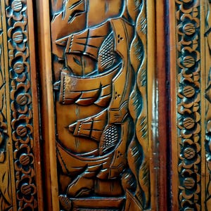May include: Close-up of a wooden panel with intricate carvings. The design features detailed depictions of sailing ships, waves, and floral patterns. The wood has a warm, brown tone with darker accents, showcasing the craftsmanship.