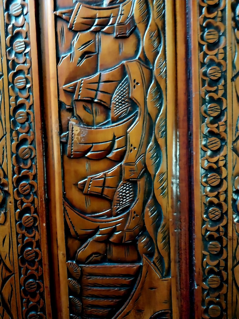 May include: Close-up of a wooden panel with intricate carvings. The design features detailed depictions of sailing ships, waves, and floral patterns. The wood has a warm, brown tone with darker accents, showcasing the craftsmanship.