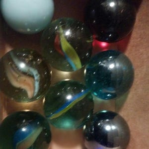 May include: A collection of marbles in various colors and patterns. The marbles include a light blue, black, and several clear ones with colorful swirls of blue, green, yellow, and red. They are contained in a brown box.