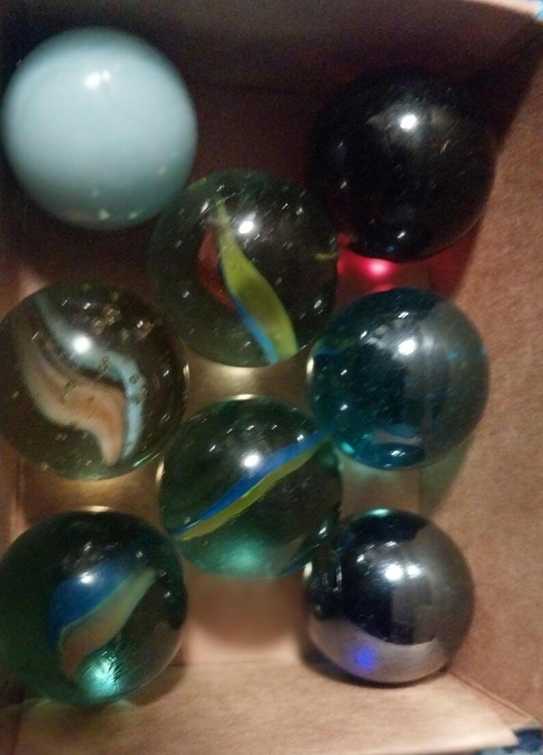 May include: A collection of marbles in various colors and patterns. The marbles include a light blue, black, and several clear ones with colorful swirls of blue, green, yellow, and red. They are contained in a brown box.