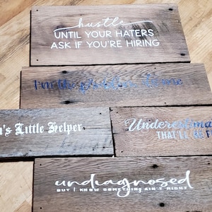 May include: A collection of rustic wooden signs with various white and blue text phrases. The signs include phrases like "hustle," "Karma's Little Helper," and "Undermate me that it'll be fun." The signs are made of weathered wood.