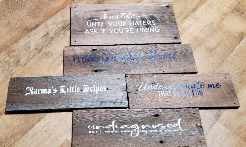 May include: A collection of rustic wooden signs with various white and blue text phrases. The signs include phrases like "hustle," "Karma's Little Helper," and "Undermate me that it'll be fun." The signs are made of weathered wood.