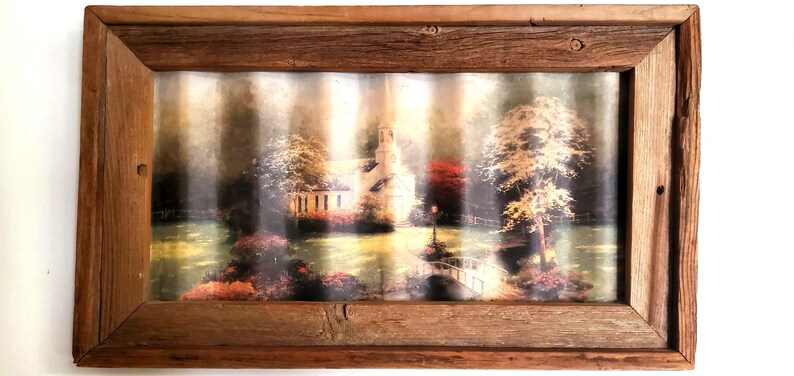 May include: A rustic wooden framed art print featuring a serene landscape with a church, bridge, and trees. The artwork has a vintage aesthetic, with soft colors and a textured appearance. The frame is made of weathered wood.