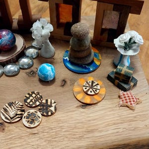 May include: A collection of decorative objects on a wooden surface. Includes wooden structures, stacked stones, floral arrangements, painted spheres, and laser-cut wooden circles with geometric designs. The color palette features natural wood tones, blues, and whites.