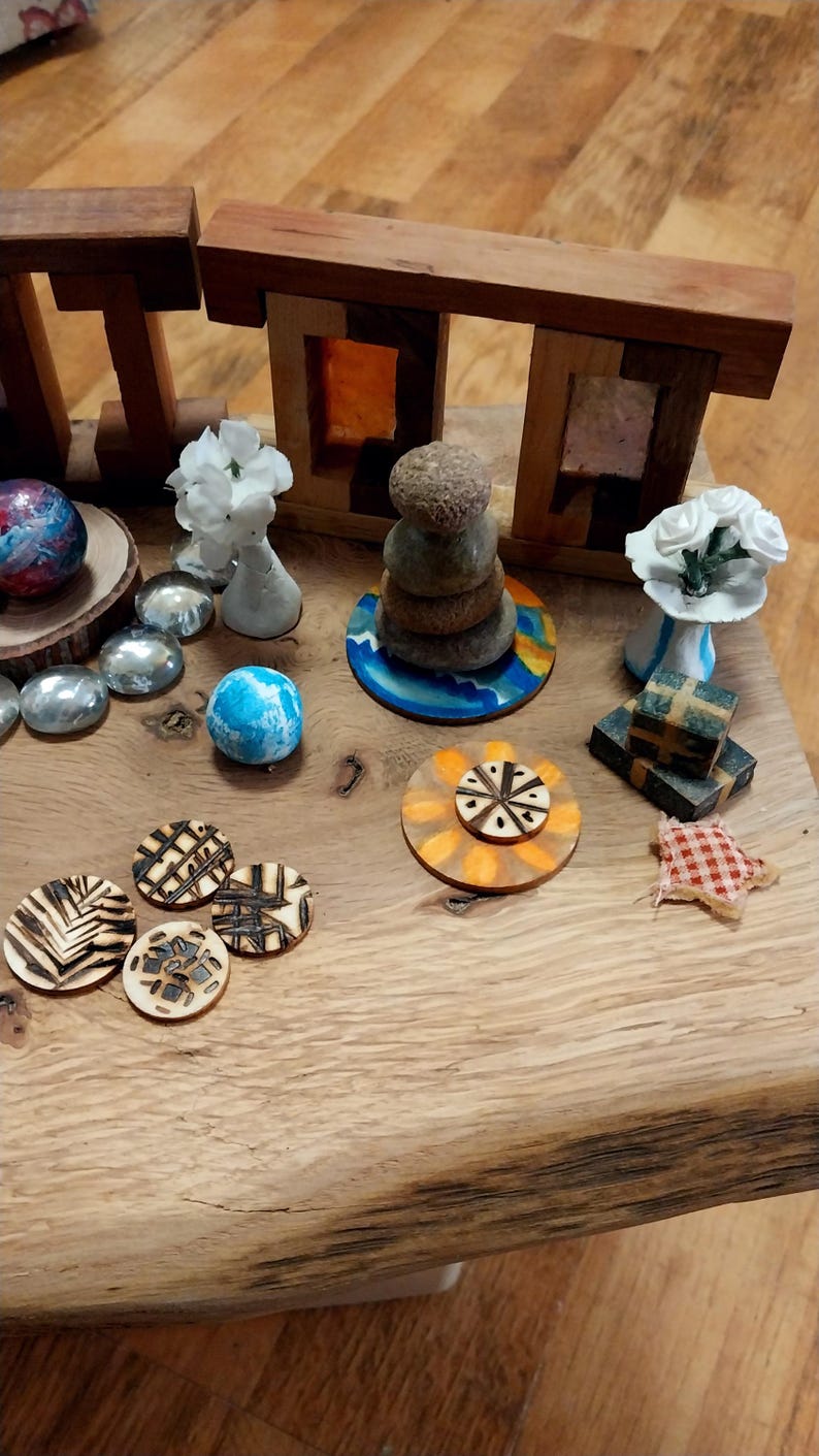 May include: A collection of decorative objects on a wooden surface. Includes wooden structures, stacked stones, floral arrangements, painted spheres, and laser-cut wooden circles with geometric designs. The color palette features natural wood tones, blues, and whites.