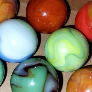 May include: A collection of colorful glass marbles in various colors, including red, orange, green, blue, and yellow. The marbles have swirled patterns and are displayed in a cardboard box. These marbles are a classic toy.