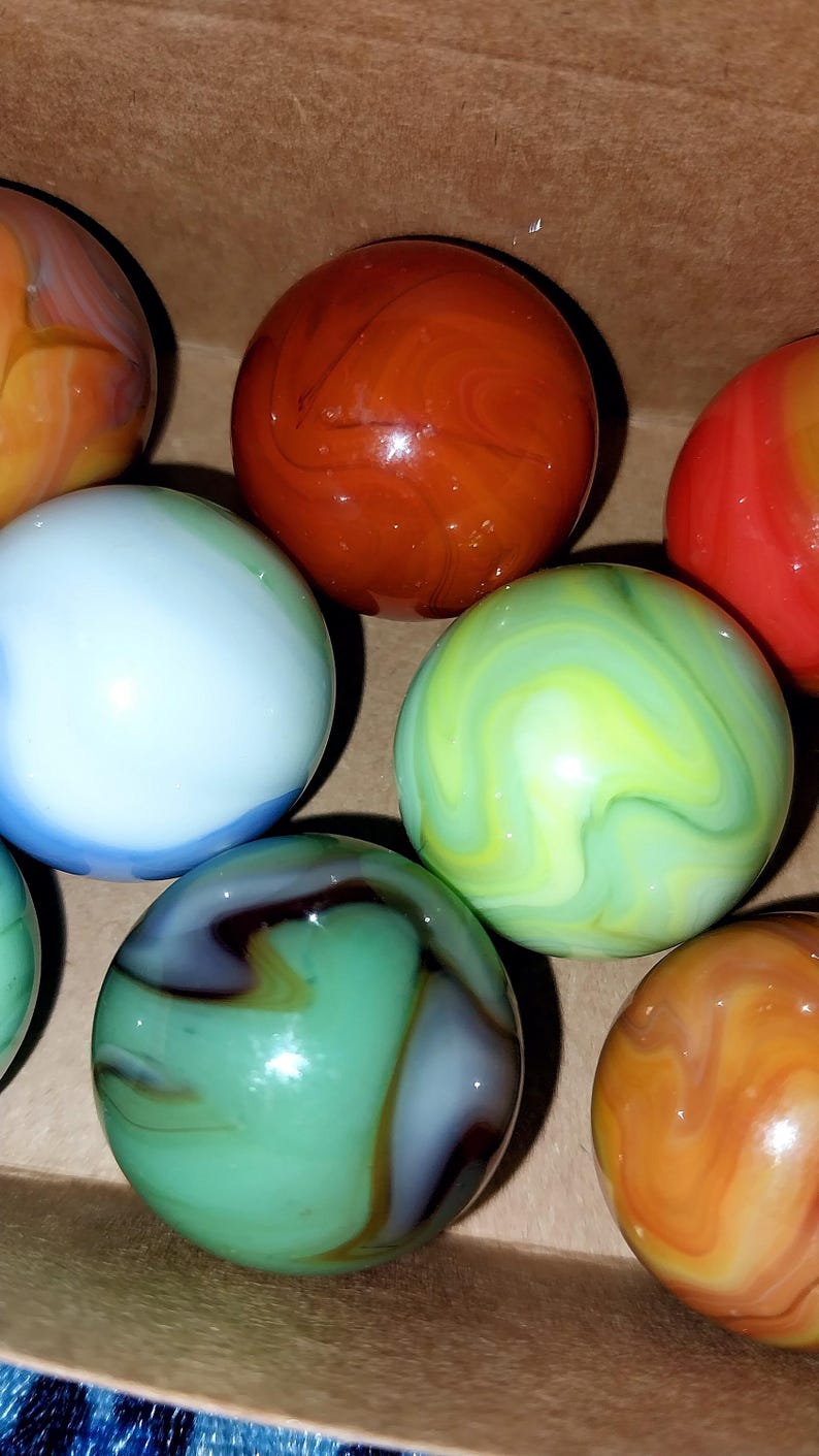 May include: A collection of colorful glass marbles in various colors, including red, orange, green, blue, and yellow. The marbles have swirled patterns and are displayed in a cardboard box. These marbles are a classic toy.