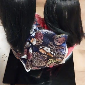 May include: A Japanese doll with long black hair and a colorful kimono. The kimono features a navy blue background with floral and geometric patterns. The doll is displayed on a black rectangular base.