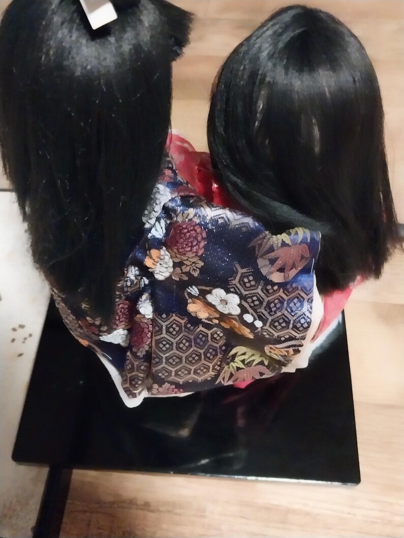 May include: A Japanese doll with long black hair and a colorful kimono. The kimono features a navy blue background with floral and geometric patterns. The doll is displayed on a black rectangular base.