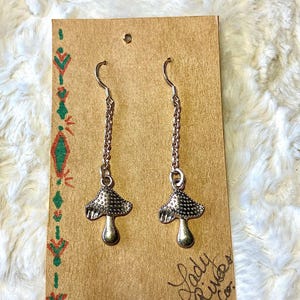 May include: A pair of silver mushroom earrings suspended from chains. The earrings are presented on a brown card with green and red decorative patterns and the text "Lady River Co."