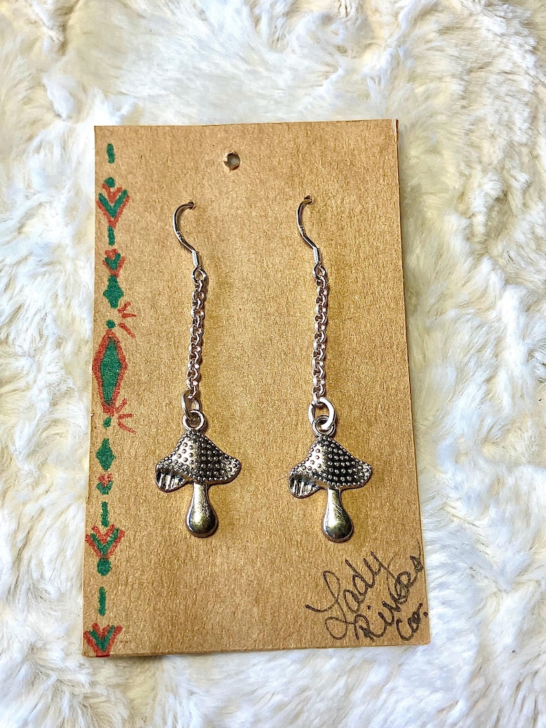 May include: A pair of silver mushroom earrings suspended from chains. The earrings are presented on a brown card with green and red decorative patterns and the text "Lady River Co."