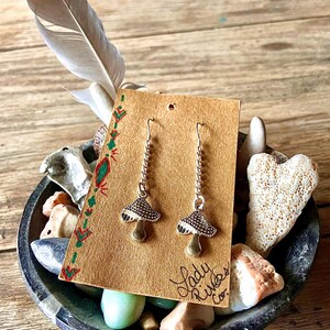 May include: Silver-coloured mushroom earrings with detailed caps and long chains, presented on a brown card with green and red decorations. The card is in a decorative bowl with stones and a feather.