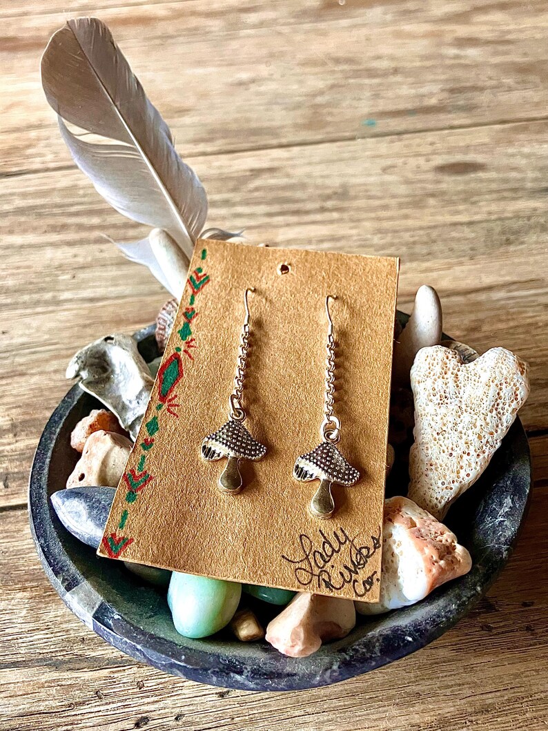 May include: Silver-coloured mushroom earrings with detailed caps and long chains, presented on a brown card with green and red decorations. The card is in a decorative bowl with stones and a feather.