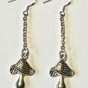 May include: A pair of silver mushroom earrings. Each earring has a mushroom charm hanging from a chain and a hook. The mushroom caps have a textured surface, and the stems are smooth.