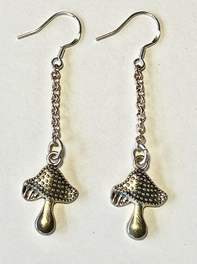 May include: A pair of silver mushroom earrings. Each earring has a mushroom charm hanging from a chain and a hook. The mushroom caps have a textured surface, and the stems are smooth.
