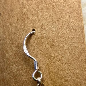 May include: Close-up of a silver earring hook with a coiled wire design. The hook is attached to a small, round, silver ring. The earring is displayed on a brown cardboard backing.