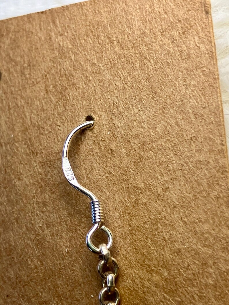 May include: Close-up of a silver earring hook with a coiled wire design. The hook is attached to a small, round, silver ring. The earring is displayed on a brown cardboard backing.