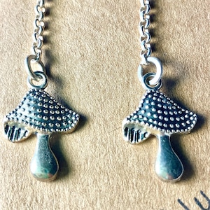 May include: Silver-coloured mushroom earrings. Each earring has a detailed mushroom cap with a dotted pattern and a smooth stem. The earrings hang from delicate chain links, creating a whimsical accessory.