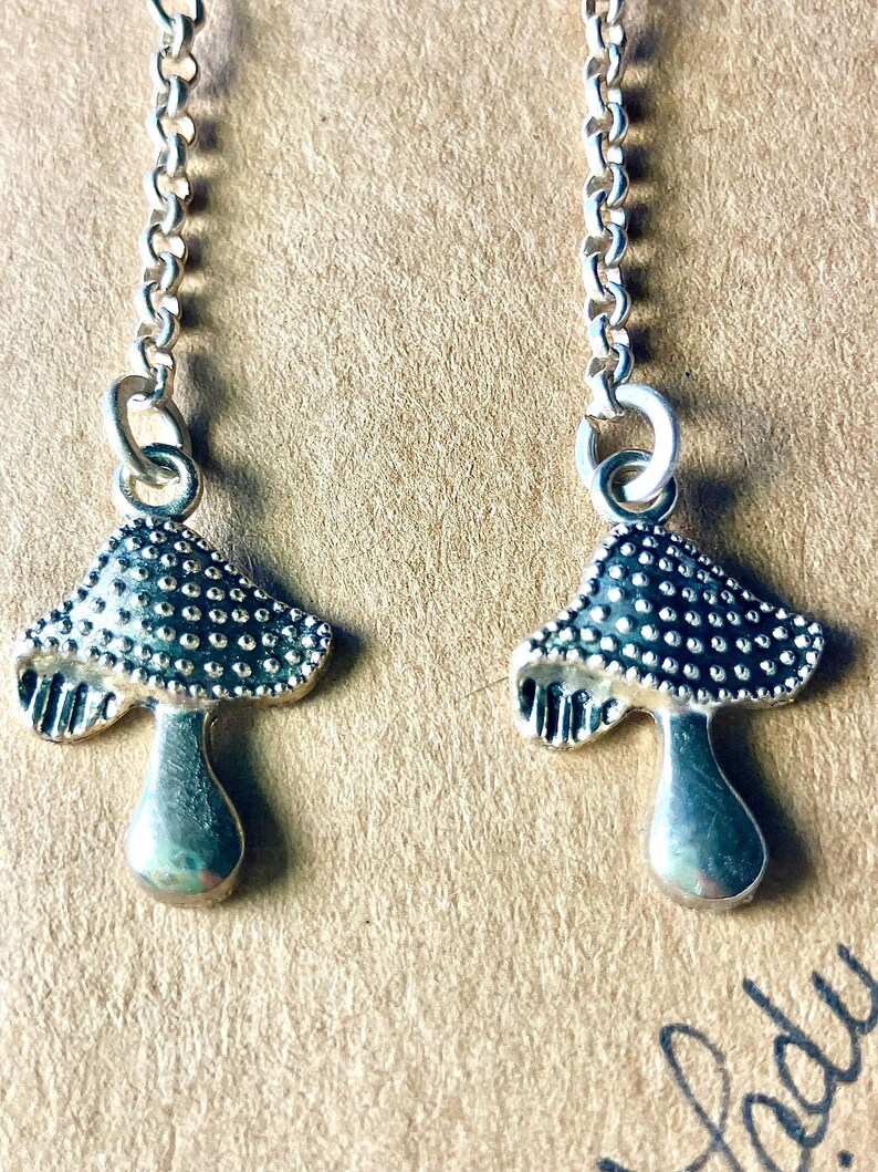 May include: Silver-coloured mushroom earrings. Each earring has a detailed mushroom cap with a dotted pattern and a smooth stem. The earrings hang from delicate chain links, creating a whimsical accessory.