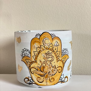 May include: White ceramic planter with a gold hamsa hand design. The hamsa hand features a lotus flower and the Om symbol in the center.