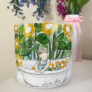 May include: White ceramic planter with a hand-painted design of sunflowers and a child in a bathtub. The planter features the name "Luca Ace" in script. The background includes green leaves and yellow flowers.