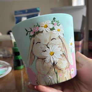 May include: A light blue ceramic planter with a hand-painted design of a white rabbit wearing a daisy flower crown and holding a daisy. The rabbit is surrounded by green leaves and white daisies.