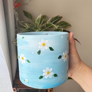 May include: A light blue ceramic planter with a white ombre effect and hand-painted white daisies with yellow centers.