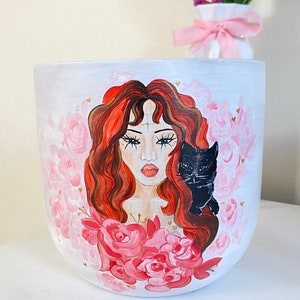 May include: White ceramic planter with a hand-painted design. The design features a woman with red hair, surrounded by pink roses and a black cat. Gold accents are scattered throughout the design. The planter is a decorative item.