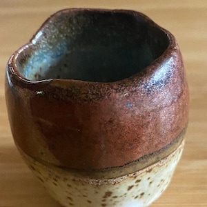 Op de afbeelding: Een klein, handgemaakt keramiek kopje met een bruine, witte en beige glazuur. Het kopje heeft een onregelmatige vorm en een licht getextureerd oppervlak.