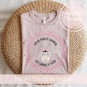 Könnte beinhalten: Hellrosa T-Shirt mit einem Cartoon-Esel mit rosa Schleife, umgeben von einem sternenbesetzten rosa Kreis mit dem Text "Con mi Burrito Sabanero Voy Camino de Belén". Das T-Shirt liegt auf einer hellbraunen, gewebten Oberfläche.