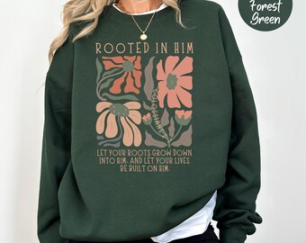 May include: Forest green sweatshirt with the text "ROOTED IN HIM" and a floral design in peach and brown. The text "COLOSSIANS 2" is also visible. The bottom of the sweatshirt has the text "LET YOUR ROOTS GROW DOWN INTO HIM. AND LET YOUR LIVES BE BUILT ON HIM."