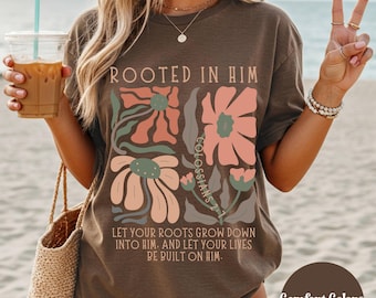 May include: Espresso-coloured t-shirt featuring a floral design and the text "ROOTED IN HIM." The design includes flowers in shades of peach, pink, and green. The text "LET YOUR ROOTS GROW DOWN INTO HIM. AND LET YOUR LIVES BE BUILT ON HIM." is also printed on the shirt.