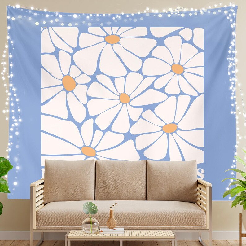 May include: A blue and white tapestry with a floral pattern of white daisies with orange centers. The tapestry is hanging on a wall above a light brown couch.