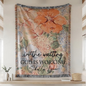 May include: A woven throw blanket featuring a watercolor-style floral pattern in shades of orange, blue, and green. The text "in the waiting GOD IS WORKING" and the name "Shelly Lynn" are printed in black script and sans-serif lettering.