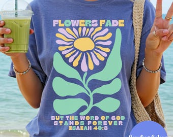 May include: A blue Comfort Colors t-shirt with a floral design and the text "FLOWERS FADE BUT THE WORD OF GOD STANDS FOREVER ISAIAH 40:8". The graphic features a large flower in yellow, purple, and green. The shirt is a Flo Blue colour.