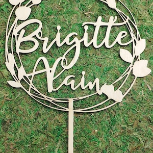 Op de afbeelding: Een houten taarttopper met een rond ontwerp met een krans van bladeren en de namen "Brigitte" en "Alain" in cursief schrift.