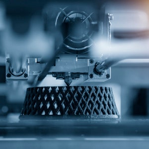 Peut inclure: Gros plan sur une imprimante 3D en fonctionnement, avec un objet en plastique noir en cours d'impression. La buse de l'imprimante est visible, extrudant du filament de plastique pour créer un motif en treillis.