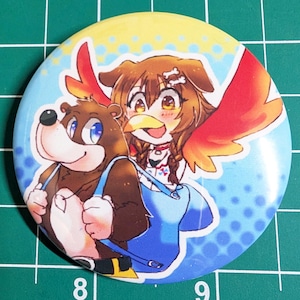 May include: A round button featuring a cartoon illustration of a bear and a character with dog ears and bird wings. The button has a blue and yellow background with a gradient effect. The bear is brown, and the other character has brown hair and red wings.
