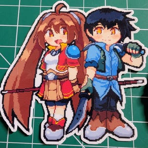 Puede incluir: Una pegatina con dos personajes de pixel art. Uno tiene pelo largo castaño, un atuendo rojo y blanco y una lanza. El otro tiene pelo negro, un atuendo azul y una espada. La pegatina está sobre una superficie verde.