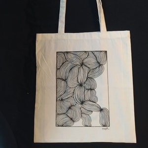 Peut inclure: Sac fourre-tout en toile blanche avec un motif abstrait en noir et blanc. Le motif est dans un cadre rectangulaire et le mot "mefi" est écrit sous le cadre.