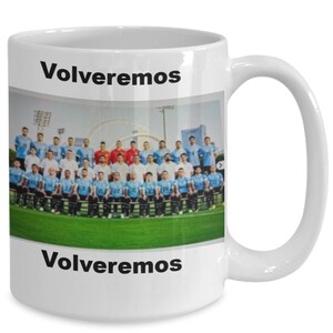 May include: White ceramic coffee mug with a photo of a soccer team in blue and white uniforms. The text "Volvereemos" is printed on the mug in black.