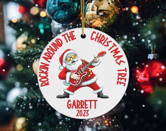 May include: A white round Christmas ornament with a red and white illustration of Santa Claus playing a red electric guitar. The text "Rockin' Around The Christmas Tree" is written in a red circle around Santa. The text "Garrett 2023" is written below Santa.