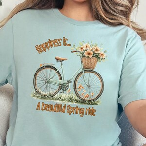 Könnte beinhalten: Ein hellblaues T-Shirt mit einem Aufdruck eines Fahrrads mit Blumen in einem Korb. Der Text auf dem Shirt lautet "Happiness is... A beautiful Spring ride."