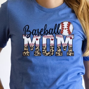 May include: A royal blue t-shirt with the text "Baseball Mom" in black lettering. The text is layered over a baseball graphic with red stitching and a leopard print pattern.