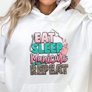 May include: White hoodie with a graphic print that says "Eat Sleep Manicure Repeat". The graphic features a pink nail polish bottle, a nail file, and a leopard print design.