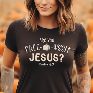 Può includere: Una maglietta nera con una grafica bianca che dice "Are you Fall-Ween Jesus? Matthew 4:19" con un'illustrazione di zucca.