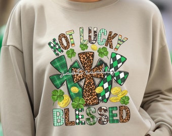 Puede incluir: Una sudadera beige con un estampado gráfico verde, negro y blanco que dice "Not Lucky Just Really Blessed". El gráfico presenta una cruz hecha de tela de estampado de leopardo con diseños de tréboles alrededor.