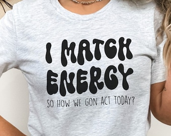 May include: Gray t-shirt with black text that reads "I match energy so how we gon act today?" The text is in a retro, wavy font.
